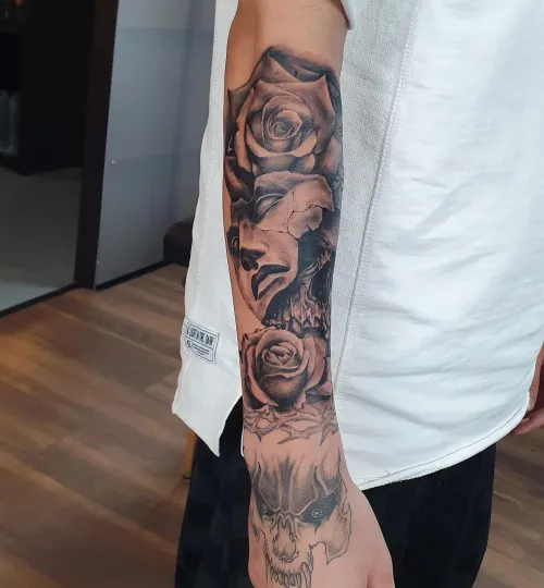 Tatuaj black and grey pe antebraț, cu trandafiri realiști, un craniu și un chip fragmentat integrat în compoziție.
