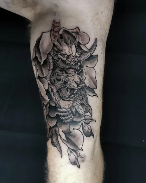 Tatuaj black and grey pe gambă, reprezentând un tigru cu armură de samurai și mască oni, integrat în elemente florale și frunze.